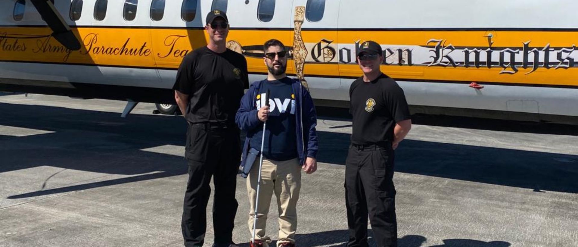Man in IBVI shirt holding a cane standing with two other men in front of a plane