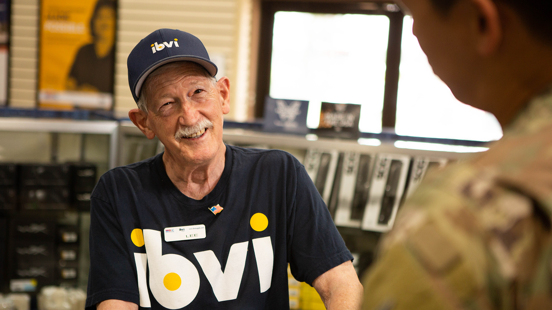 Elderly man wearing a blue IBVI shirt and hat. He has a nametag on his shirt that says “LEE” and a United States flag pin. He is assisting military personnel in a store.