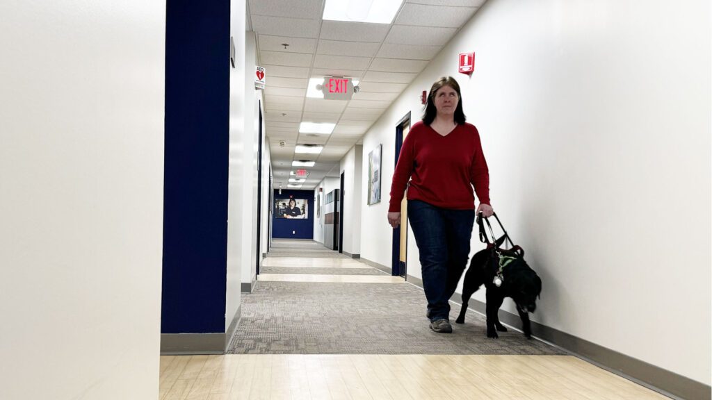 Woman walking with guide dog down a hallway with carpet and laminate floors indicating where doors are.